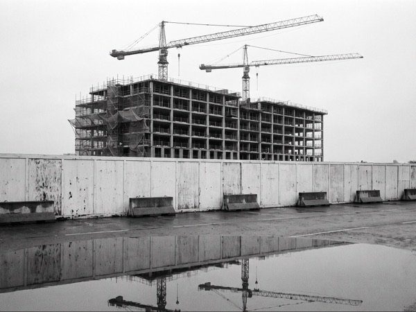Half-built concrete frame and two tower cranes rising behind white site hoarding